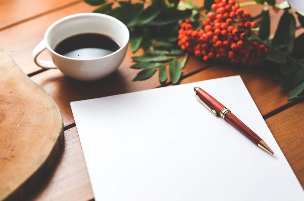 Writing pad on a table with a red ballpoint pen sitting on it. A coffee cup sits on the table next to a berry branch.